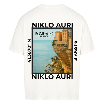 Weißes T-Shirt mit Rückendruck einer Illustration von Bonifacio, Frankreich. Zu sehen sind Häuser auf dramatisch abfallenden Felsen an der Mittelmeerküste. Der türkisfarbene Ozean erstreckt sich im Hintergrund. Oben und unten steht „NIKLO AURI“, seitlich die Koordinaten 41.3870° N und 9.1590° E. Mittig ist der Schriftzug „BONIFACIO – FRANCE“ platziert.