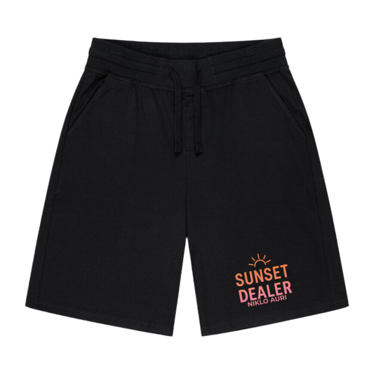 Schwarze Shorts mit seitlichen Eingrifftaschen und Kordelzug am Bund. Auf dem rechten Hosenbein befindet sich ein farbenfroher Print: Oben eine stilisierte untergehende Sonne, darunter die Schriftzüge „SUNSET DEALER“ in orange und pink sowie „NIKLO AURI“ in klein darunter. Der Druck verleiht der Hose einen urbanen, sommerlichen Look.